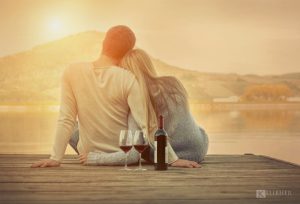 a man and woman sitting on a dock with wine glasses and a lake